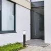 A modern house entrance with a black door, white brick wall, and a black and white bollard light in front of some greenery. Produktbild-Vorschau
