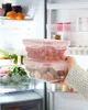 A person is holding two clear plastic food storage containers filled with cherry tomatoes and mushrooms, placing them into an open refrigerator. Produktbild-Vorschau