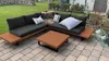 An outdoor lounge set with cushions and a coffee table on a paved patio, with a grassy area and a stone wall in the background. Produktbild-Vorschau