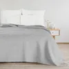 A neatly made bed with a gray blanket and white pillows, next to a small wooden nightstand with candles and a vase. Náhľad obrázku produktu
