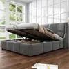A grey upholstered bed with its storage compartment open, revealing the mattress and bedding, in a well-lit room with large windows. Produktbild-Vorschau