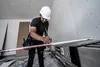 A construction worker is measuring a plank on a ladder in an unfinished room. Produktbild-Vorschau