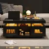 A modern living room scene featuring a black high-gloss coffee table with integrated LED lighting, a laptop, and decorative items. Náhled obrázku produktu