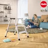 A woman is reading a magazine while sitting on the floor next to a step ladder in a living room. Náhľad obrázku produktu