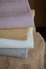 A stack of four folded blankets with a herringbone pattern, each in a different color, resting on a wooden surface. Produktbild-Vorschau