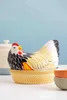 A ceramic chicken-shaped dish with a chick underneath, sitting in a yellow basket-like base. Other bowls are visible in the background. Náhled obrázku produktu