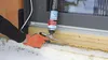 A person wearing orange gloves is using a can of spray foam to insulate around a window frame in a snowy environment. Produktbild-Vorschau