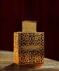 A close-up shot of a golden perfume bottle with intricate geometric patterns, placed on a wooden surface with a dark red fabric background. Produktbild-Vorschau
