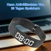 A black fitness tracker with a digital display is shown on a wooden surface in front of a laptop. Produktbild-Vorschau