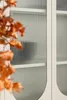 A close-up shot of a beige vitrine with fluted glass doors, partially obscured by blurred orange leaves in the foreground. Voir les images du produit