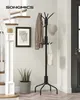A black metal coat rack with 12 hooks stands in a hallway next to a staircase and a potted plant. A shirt and a hat are hung on the coat rack, along with a tote bag. Náhled obrázku produktu