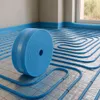 A roll of blue insulation material for underfloor heating is placed on a floor with installed heating pipes. Náhľad obrázku produktu