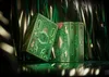 Two green playing card boxes with gold intricate designs are displayed diagonally on a reflective surface, with green and gold streamers in the background. Náhled obrázku produktu