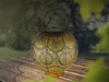 A solar-powered lantern sits on a wooden table outdoors, with a blurred green background of trees and foliage. Produktbild-Vorschau