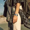 A person wearing a watch, leather jacket, and plaid shirt in an outdoor urban setting. Produktbild-Vorschau
