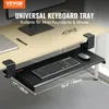 A black keyboard tray is clamped to the underside of a light wood desk, with a keyboard and mouse on it. The desk has a monitor base, a notebook, and glasses on it. Produktbild-Vorschau