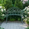 A vintage-style green metal garden bench with a rose pattern sits on a paved patio surrounded by lush green bushes and blooming pink and white roses. Podgląd zdjęcia produktu