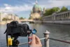 A hand inserts an SD card into a camera with a city skyline and river in the background. Podgląd zdjęcia produktu