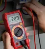 A person is using a red digital multimeter to test electrical voltage in an electrical panel. Podgląd zdjęcia produktu
