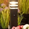 A black outdoor bollard light with a motion sensor is shown in a garden setting with plants and flowers. Produktbild-Vorschau