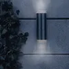 A silver outdoor wall lamp is mounted on a textured gray wall, with ivy growing on the left side. The lamp emits light upwards and downwards. Produktbild-Vorschau