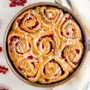 A close-up overhead view of a round pan filled with baked cinnamon rolls with a red berry filling and white icing. Produktbild-Vorschau
