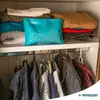 A turquoise dehumidifier bag is placed on a shelf in a closet, surrounded by folded clothes and hanging garments. Náhled obrázku produktu
