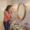 A woman is putting on earrings in front of a mirror, with a console table below holding various decorative items and accessories. Produktbild-Vorschau