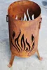 A rusty metal fire pit with flame cutouts and three legs, placed on a concrete surface. Produktbild-Vorschau