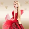 A Barbie doll wearing a red holiday dress with blonde hair and large white earrings, set against a gold background with snowflake graphics. Náhled obrázku produktu