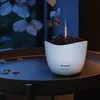A white aroma diffuser with a flame-like mist sits on a dark round table with scattered petals. Produktbild-Vorschau