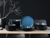 A set of blue and black ceramic dishes arranged on a wooden table with a vase and branches in the background. Náhľad obrázku produktu