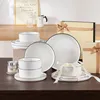 A collection of white ceramic dishes with black trim, arranged on a wooden table, with gift boxes and a decorative item in the background. Voir les images du produit