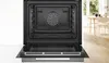 An open oven with its interior visible, set against a neutral background with a hint of a dining area in the background. Produktbild-Vorschau