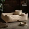 A beige corduroy dog bed with a small pillow is placed on a floor in a room with a brick wall and shelves in the background. A pet bowl is also visible. Produktbild-Vorschau
