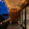 A balcony at dusk decorated with string lights, a small table and chairs, and potted plants. Produktbild-Vorschau