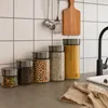 A kitchen counter with several glass storage jars filled with different food items, a wooden cutting board, and kitchen utensils hanging on the wall. Produktbild-Vorschau