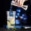 A hand is pouring liquid from a jigger into a cocktail shaker glass with recipes printed on it. Ice, lemon slices, and mint are visible inside the glass. Produktbild-Vorschau