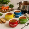 Four colorful soup bowls filled with tomato soup are arranged on a kitchen counter, with ingredients like carrots and garlic in the background and a pot of soup on a stove. Voir les images du produit