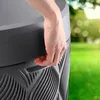 A hand is shown opening a large gray water tank in a grassy outdoor setting with blurred trees and sunlight. Náhled obrázku produktu