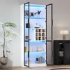 A tall, white display cabinet with glass doors and interior LED lighting, showcasing various items on its shelves. A potted plant is visible to the left, and a dark cabinet with a lamp is to the right. Voir les images du produit