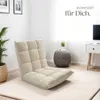 A beige floor chair is placed on a cream rug in a bright, minimalist room. A plant is visible to the left, and a small side table with a plant and a white drawer unit with a television are on the right. Náhled obrázku produktu