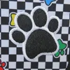 A close-up of a black and white checkered fabric with embroidered paw prints and other dog-related icons. Produktbild-Vorschau