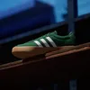 A green Adidas sneaker with white stripes and a tan sole rests on a wooden shelf. Produktbild-Vorschau