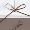 A close-up of a brown ribbon tied in a bow over a white surface with text, with a sprig of eucalyptus below. Produktbild-Vorschau