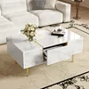 A modern white coffee table with gold accents is placed in a living room setting, next to a white sectional sofa. Produktbild-Vorschau