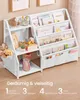 A white children's bookshelf filled with books and stuffed animals, placed against a wall with framed pictures above. Text overlays describe its features. Produktbild-Vorschau