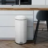 A cream-colored trash can with a black rim and pedal is placed on a wooden floor next to a white kitchen counter and a black stool. Produktbild-Vorschau