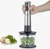 A hand holding a silver immersion blender over a clear container filled with chopped green vegetables and onions. A few onions and parsley are visible to the left. Produktbild-Vorschau