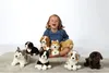 A young girl is sitting on a rug surrounded by several plush toy dogs. Produktbild-Vorschau